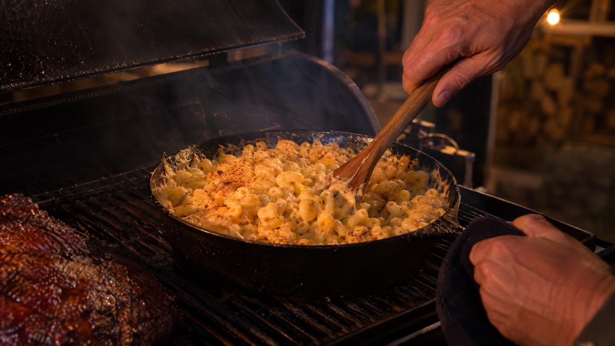 A wooden spoon stirring macaroni and cheese in a skillet on a smoker grate
