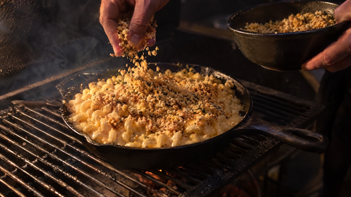 Buttered breadcrumbs being scattered over macaroni and cheese on a smoker grate