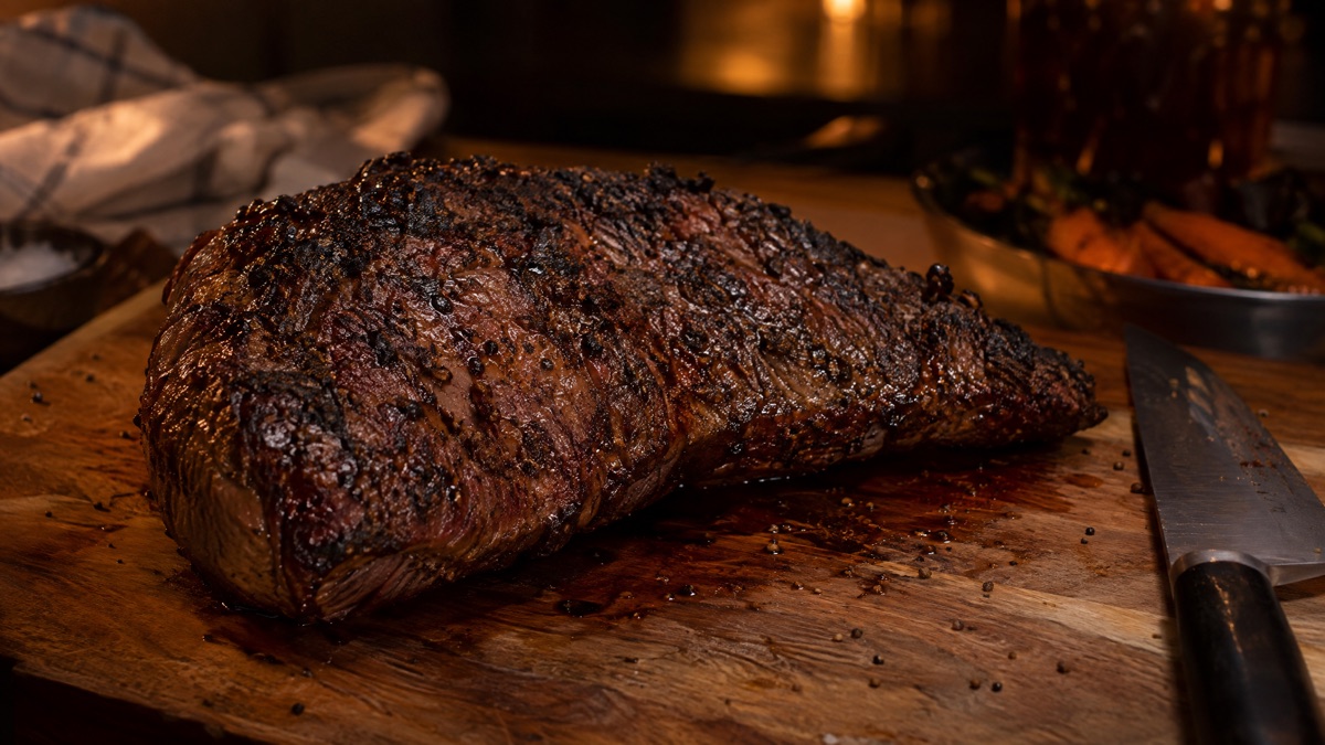 A seared tri-tip resting whole on a wooden board beside a knife