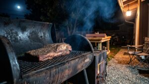 A seasoned whole packer brisket on the grate of a backyard offset smoker at night, thin blue smoke drifting toward a porch light, an empty camp chair beside the fence